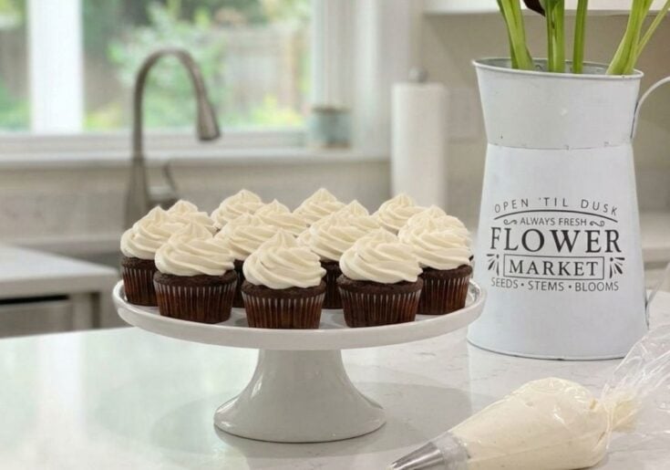 A white cake stand holding several chocolate cupcakes topped with a thick, swirl of fluffy vanilla frosting made without powdered sugar, sitting on a white kitchen counter.