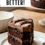 A rich, two-layer chocolate cake with swirls of buttercream frosting on a wooden stand, on a rustic kitchen table.