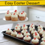Multiple rows of cupcakes with white frosting and red jelly bean toppers arranged on a rustic table with tulips in the background.