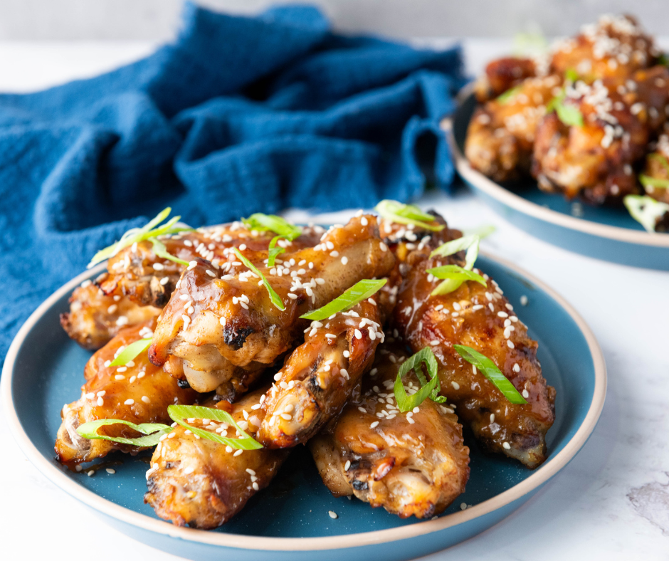 A side-angle eye-level photograph of a generous serving of glazed Chinese Chicken Wings on a round blue plate, with a second full plate blurred in the background, near a blue napkin.