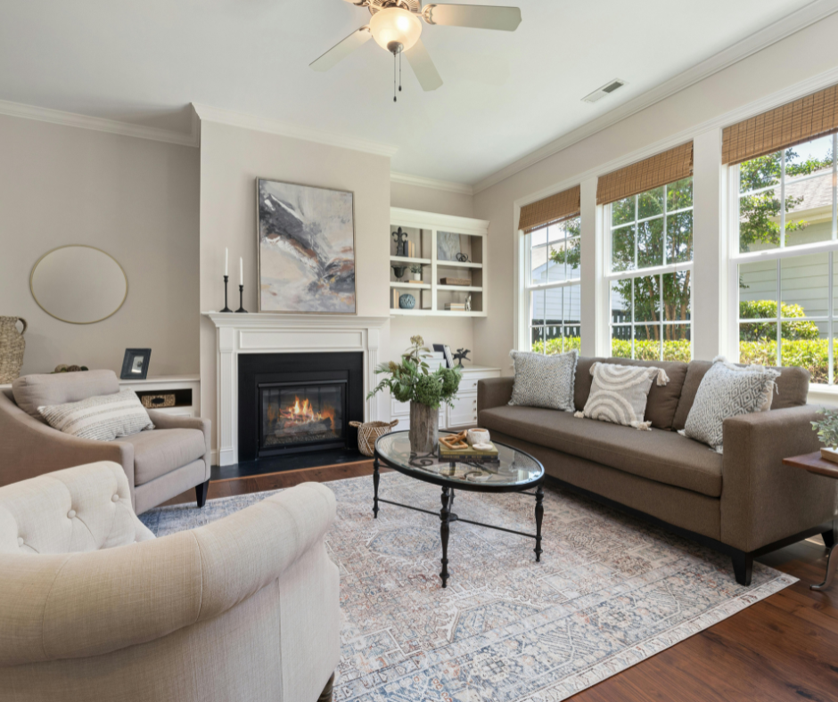 A cozy, traditional-modern living room featuring a gray sofa with patterned throw pillows, two neutral armchairs, and a black fireplace with a white mantel. A large abstract painting hangs above the fireplace, and sunlight streams through three large windows with bamboo shades.
