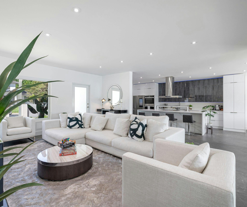 A spacious, bright open-concept living room and kitchen. The space features a large white L-shaped sectional sofa, a round marble coffee table, and minimalist white walls. In the background, a modern kitchen with dark cabinetry and a white island is visible.