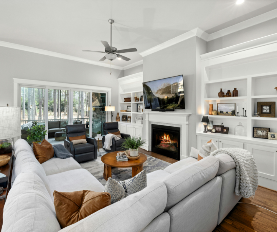 A warm, transitional living room with built-in white bookshelves, a fireplace with a lit fire, and a large wall-mounted TV. The room is furnished with a white sectional decorated with cognac-colored velvet pillows and two dark leather recliners.