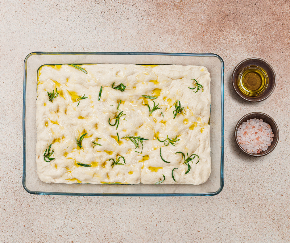 Raw focaccia dough with deep dimples, olive oil, fresh rosemary, and sea salt in a glass pan.