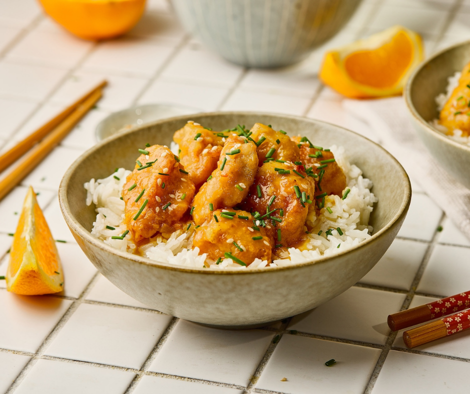 A bowl of orange chicken and white rice garnished with green onions and sesame seeds, surrounded by fresh orange wedges.