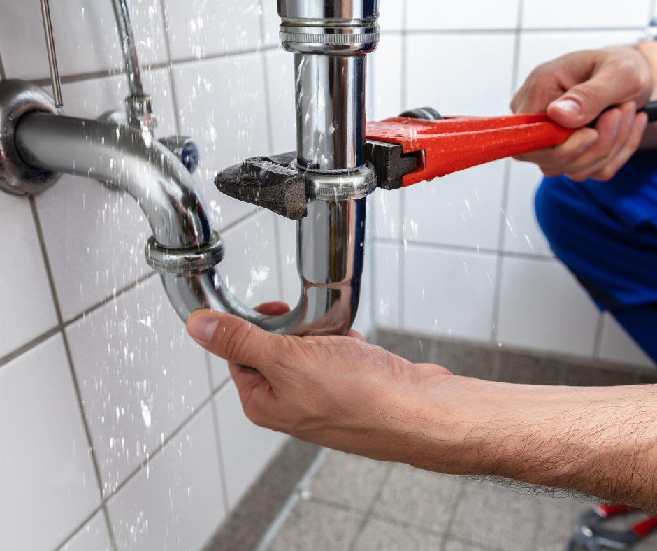 A plumber using a wrench to repair a leaky chrome P-trap pipe under a sink.