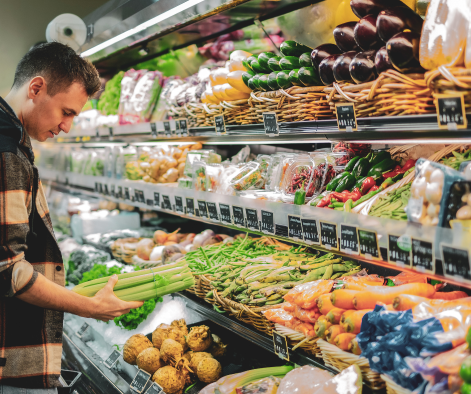 A man in a plaid jacket selecting fresh celery from a vibrant, well-stocked produce section in a grocery store.