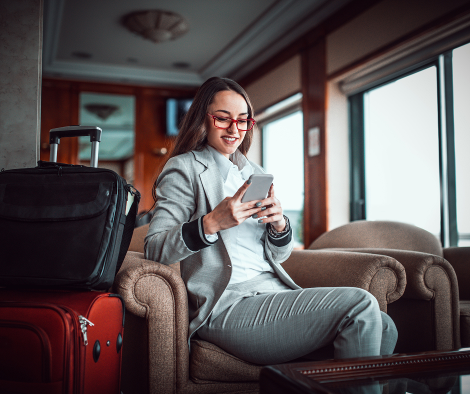 A woman in a grey suit and red glasses sitting in a lounge and looking at her smartphone.
