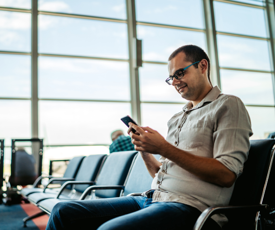 A man with glasses sitting in an airport terminal using his smartphone.