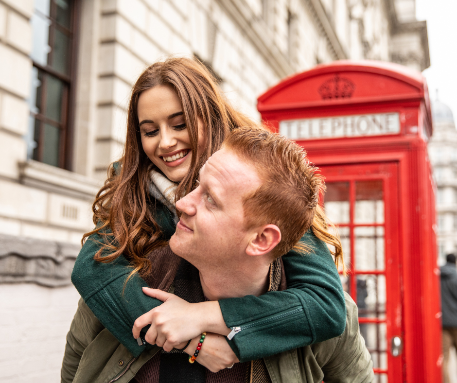 A happy man giving a woman a piggyback ride in front of a classic red British telephone box in London, depicting a successful international reunion.
