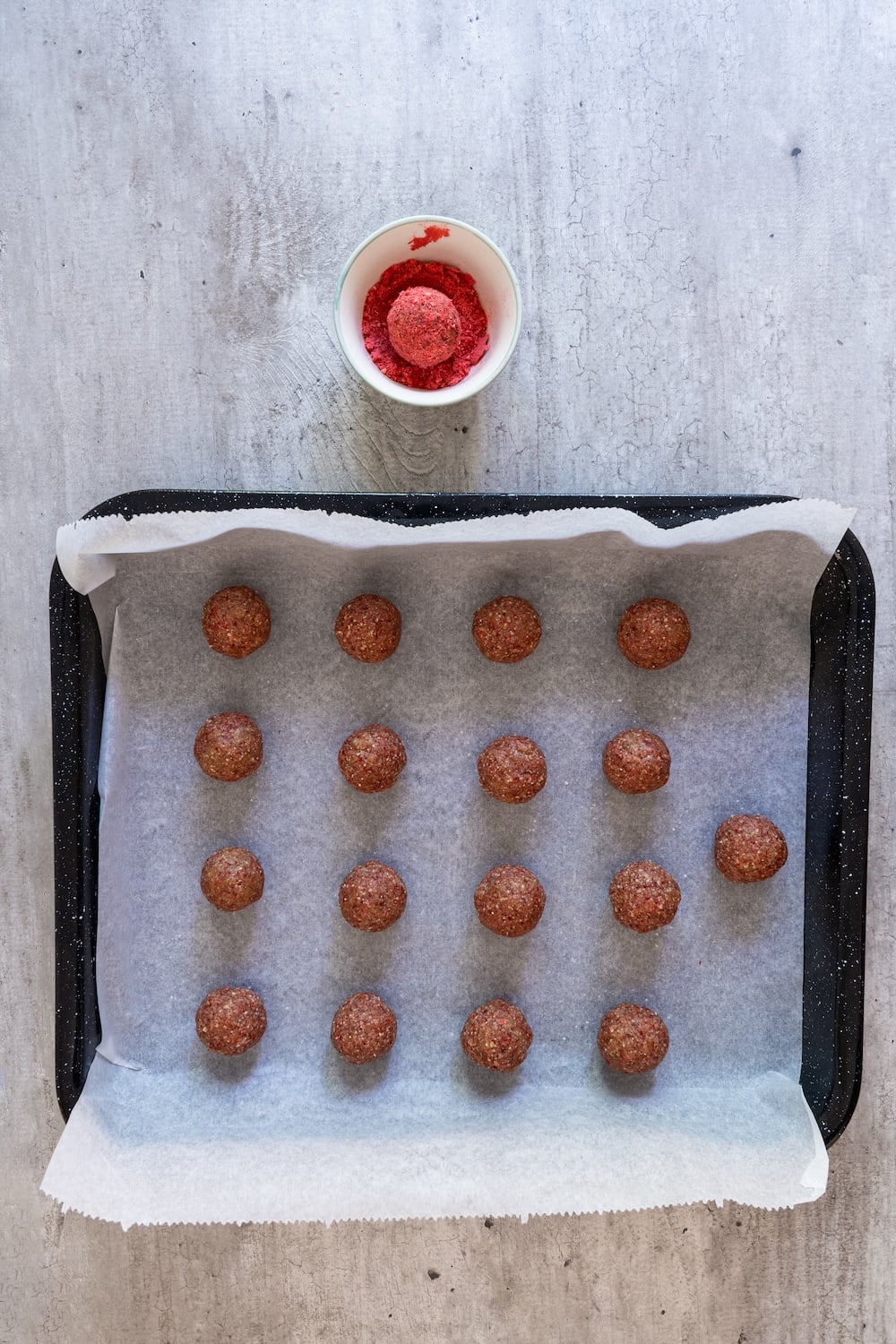 A hand rolling a single energy ball in a small bowl of bright red freeze-dried strawberry dust.