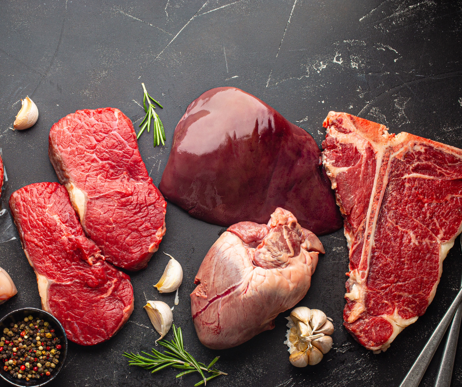 Raw beef steaks, a large piece of liver, and a whole beef heart arranged on a dark stone surface with garlic and rosemary.