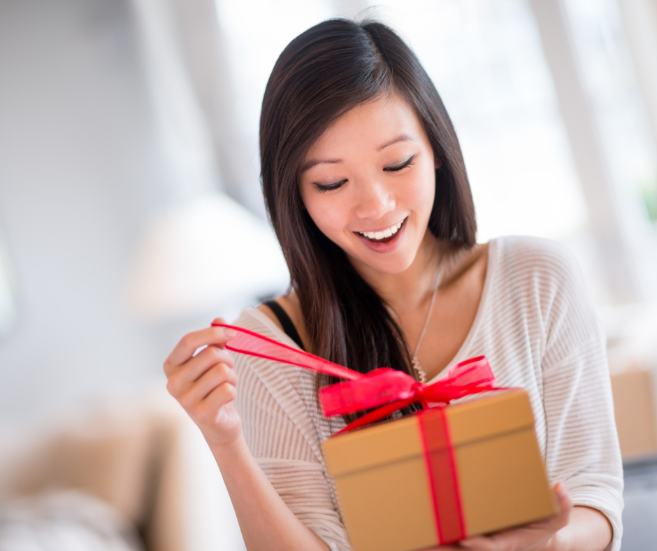 A smiling woman opening a gold gift box with a bright red ribbon in a bright, indoor setting.