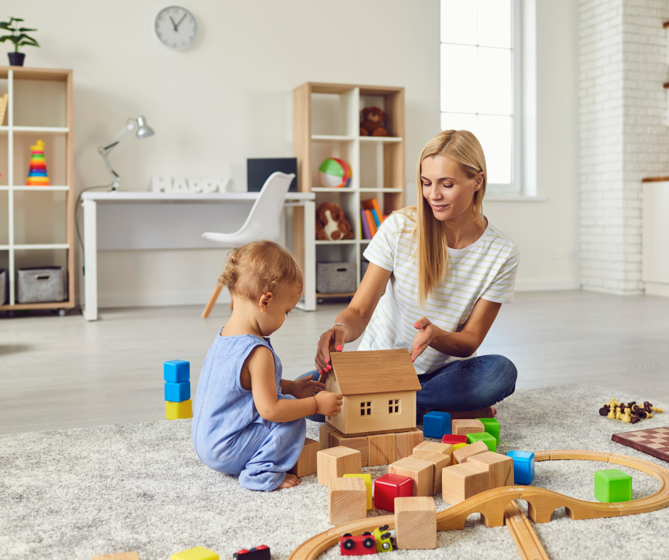 A blonde au pair sitting on a rug and helping a toddler build a house with wooden blocks in a bright playroom.