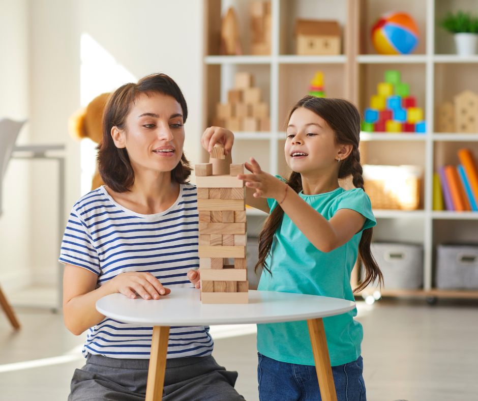 An au pair in a striped shirt playing a wooden block stacking game with a young girl at a small white table.