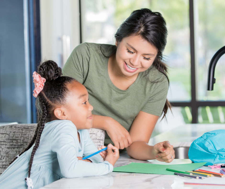 A smiling au pair helping a young girl with an art project and drawing at a kitchen counter.