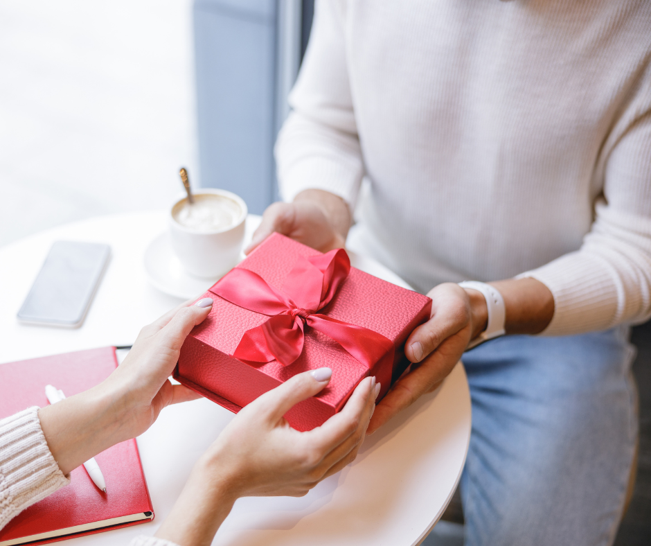 Two people sitting at a white table exchanging a textured red gift box tied with a satin red bow.