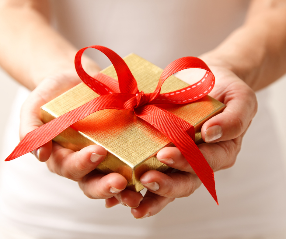 A close-up view of a person’s hands holding a small square gold gift box tied with a red ribbon.