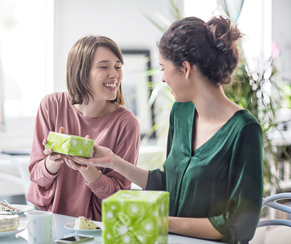 wo women laughing and sitting at a table while exchanging gifts wrapped in bright green patterned paper.