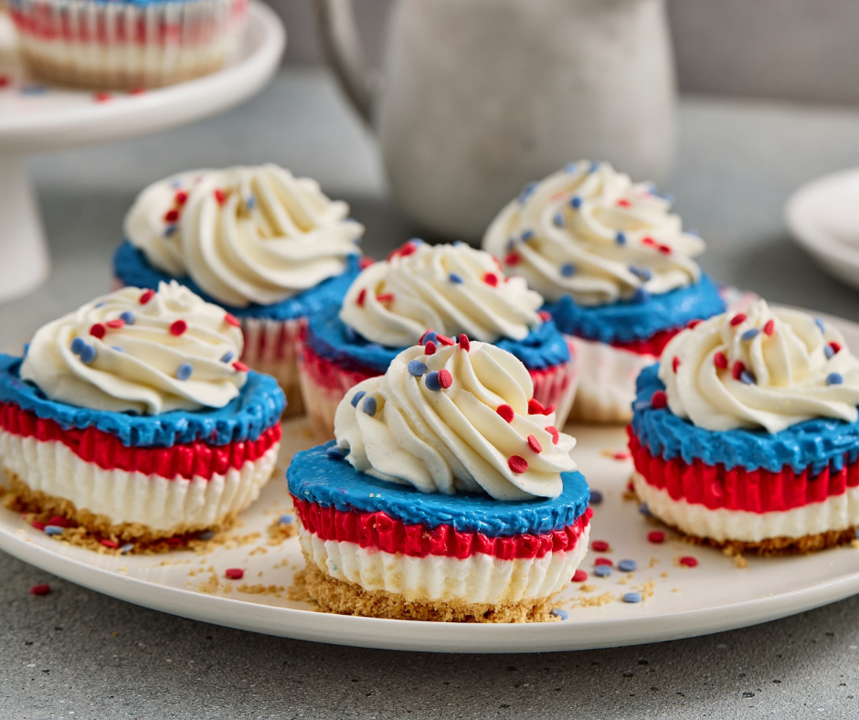Layered patriotic mini cheesecakes in paper liners displayed on a white cupcake stand, showing the distinct red, white, and blue colors for a festive holiday dessert table.