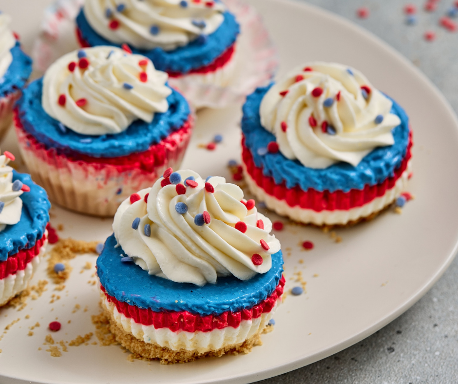 Close-up vertical view of a patriotic mini cheesecake with vibrant red and blue layers, piled high with a perfect swirl of whipped cream and red and blue star sprinkles.