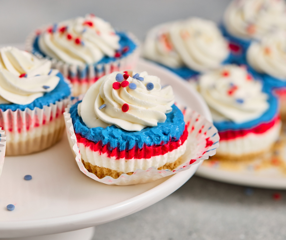 A platter of festive 4th of July mini no-bake cheesecakes made with Philadelphia cheesecake mix, showing layers of graham cracker crust, white cheesecake, red filling, and blue topping.