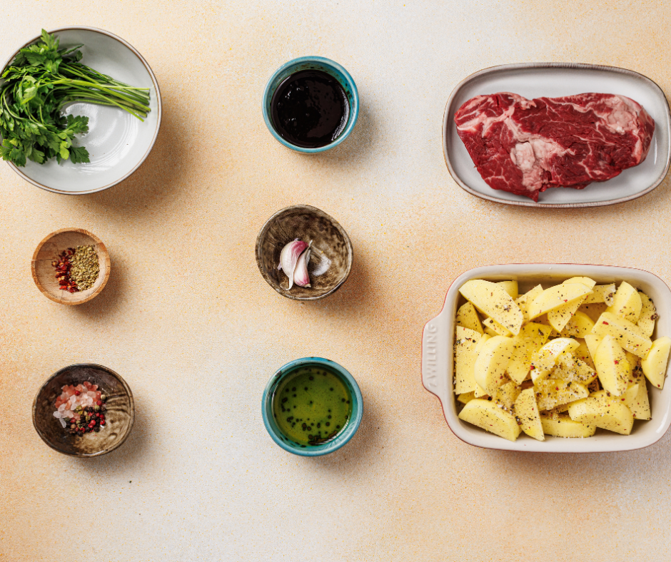 Raw ribeye steak, sliced potatoes, fresh parsley, garlic cloves, and red pepper flakes arranged for meal prep.