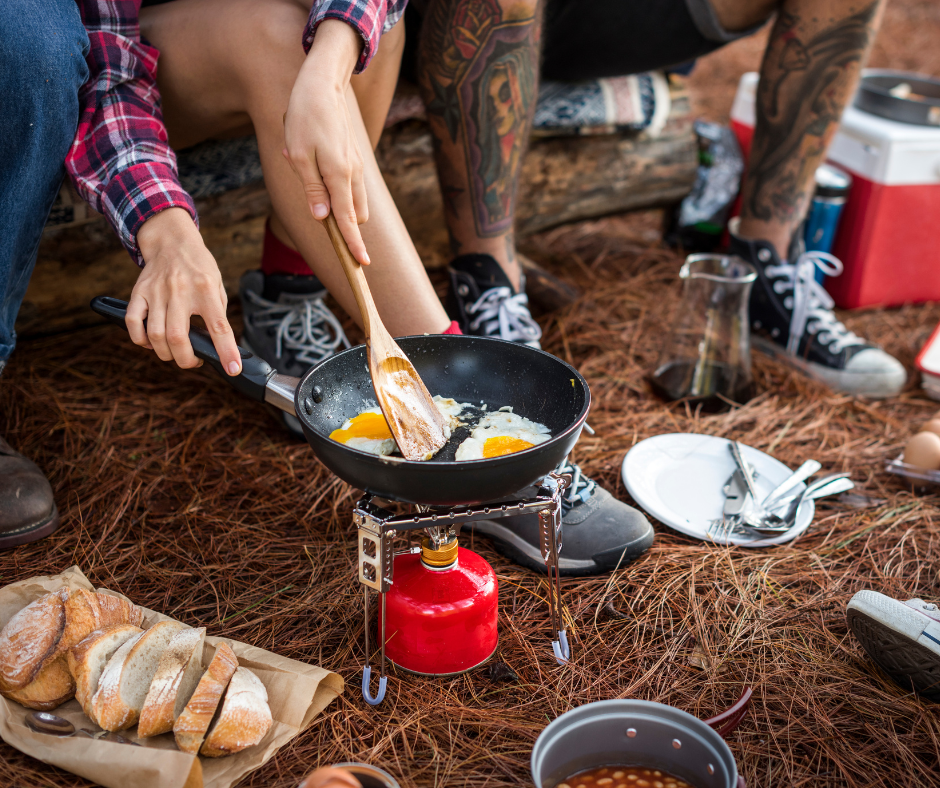Close-up of a person frying eggs in a pan over a portable camping stove on a bed of pine needles, with sliced bread and camping gear nearby.