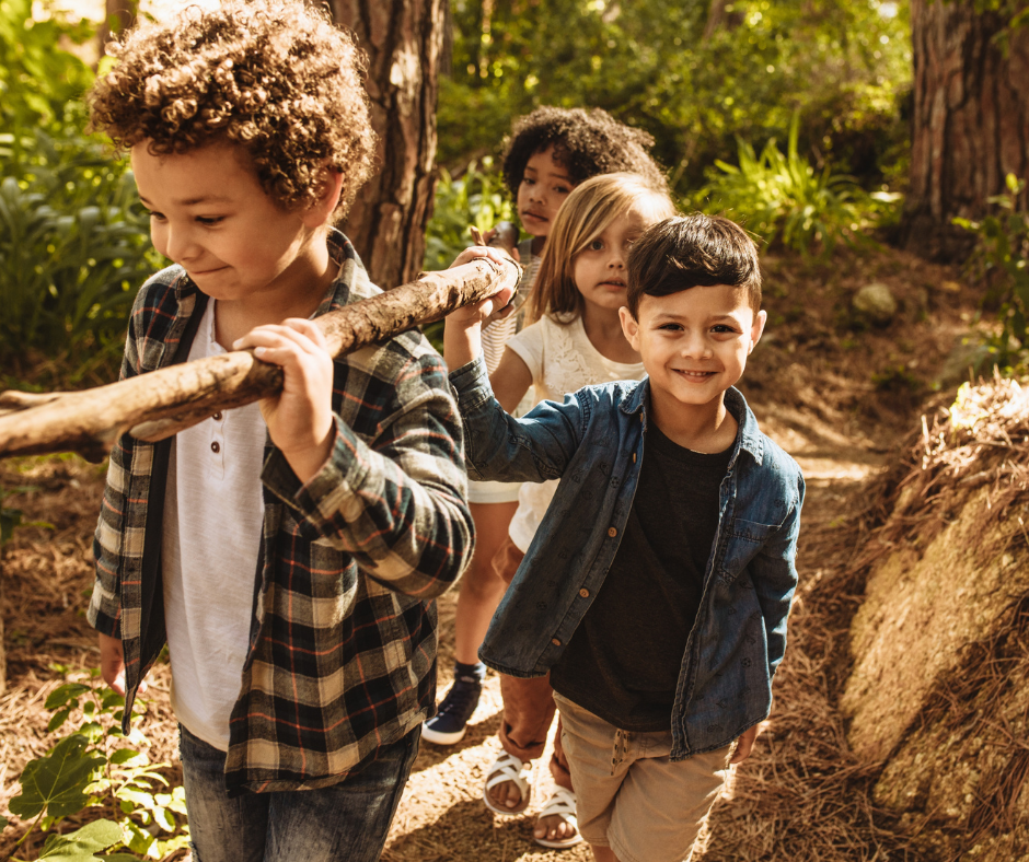 A group of four happy children playing together and carrying a large fallen branch while exploring a wooded trail during a family camping trip.