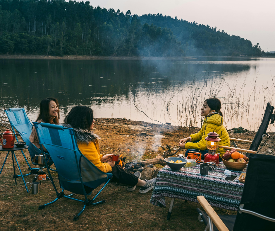 A mother and two children sitting in blue camp chairs around a small campfire by a lake at dusk with a lantern and snacks on a table.