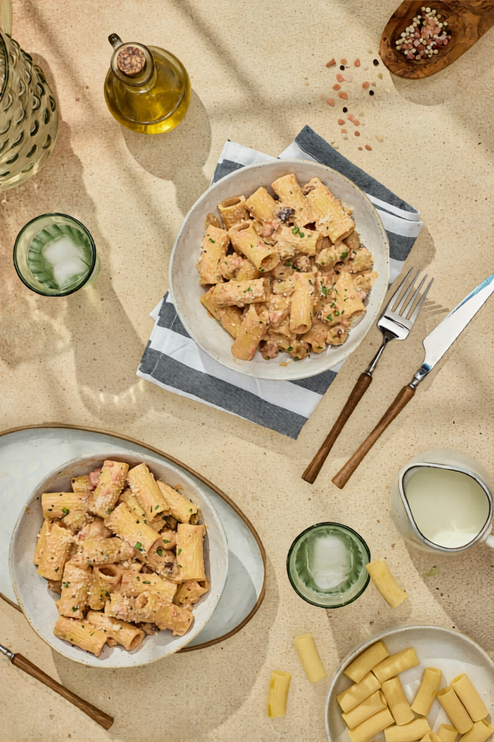 A lifestyle table setting with two bowls of rigatoni pasta, olive oil, and cream, showing a complete pasta dinner for the family.