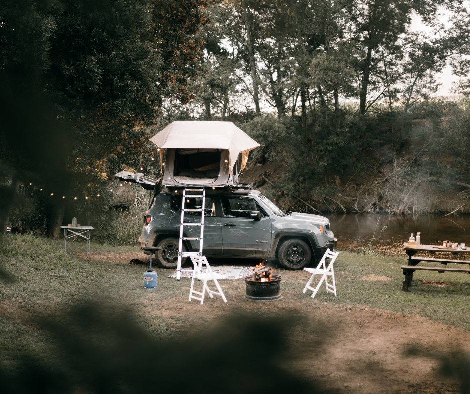 A grey SUV parked by a river at sunset featuring a rooftop tent deployed with a ladder, campfire, and string lights.