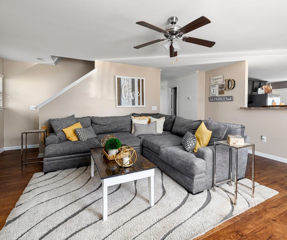 A spacious family room layout with a large gray sectional couch, a dark wood coffee table with a white base, a patterned area rug, and a dark wood ceiling fan spinning overhead.