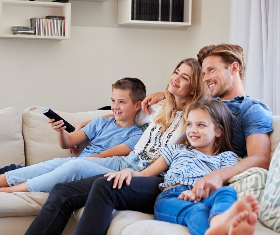 A happy family of four with a mother, father, son, and daughter sitting closely together on a comfortable couch while watching television, with the son holding a remote control.