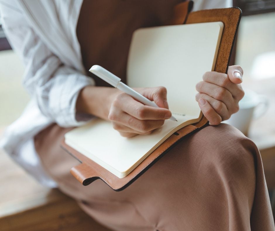 A close-up of a person sitting and writing with a white pen in a leather-bound notebook with blank pages.