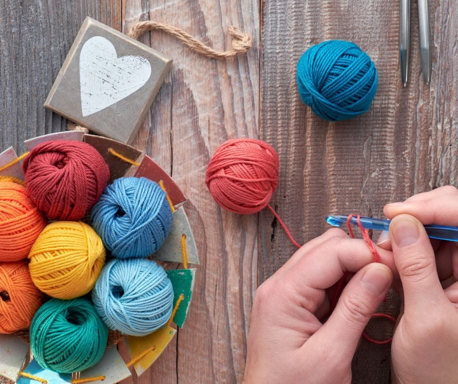 A top-down view of hands crocheting with pink yarn on a wooden table, next to a basket filled with colorful yarn balls