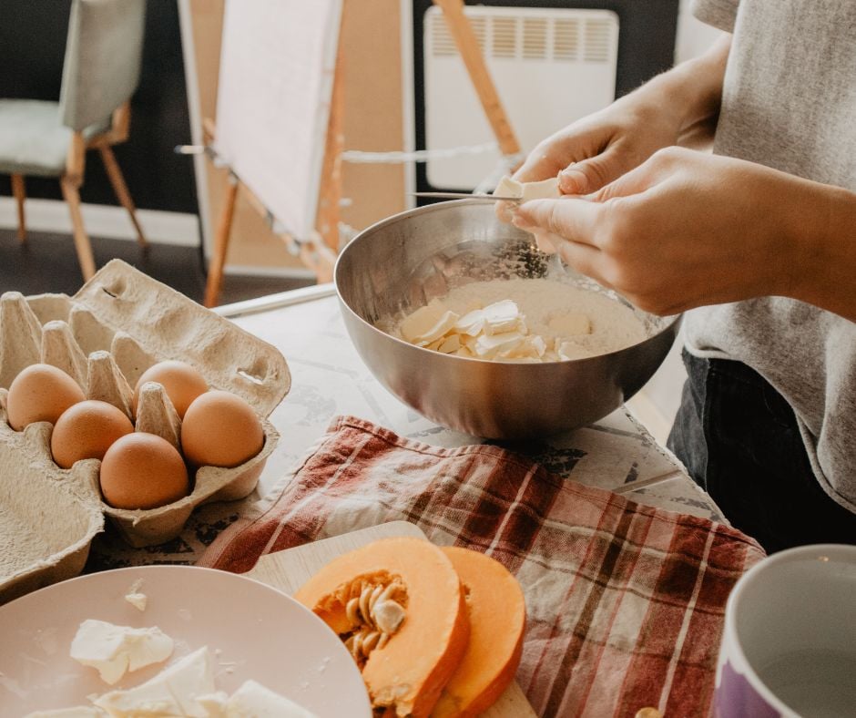 A person adding pieces of butter into a metal mixing bowl with flour, with a carton of eggs and sliced pumpkin on the kitchen counter.