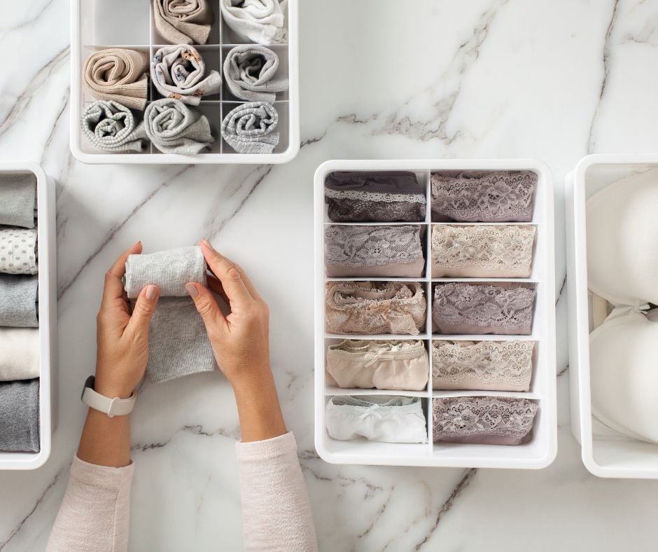 Overhead view of a person neatly rolling a grey cotton sock next to drawer organizers filled with folded lace underwear and bras on a marble countertop.
