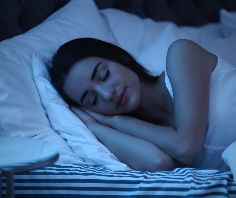 A woman sleeping peacefully in bed at night, resting her head on a white pillow under soft blue lighting.