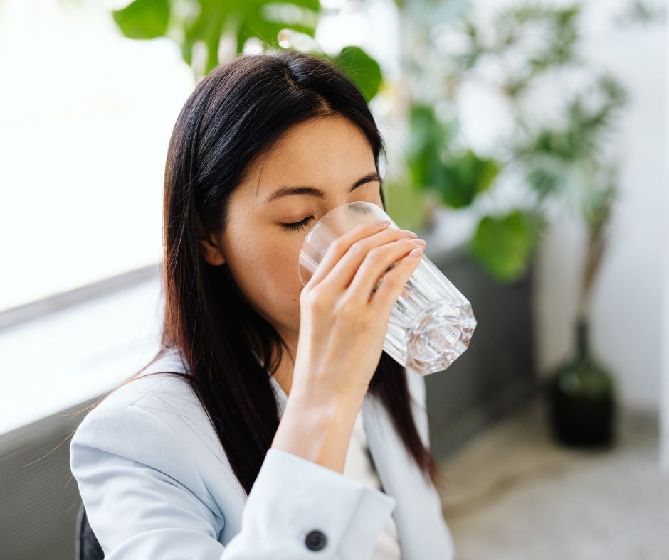 A close-up of a woman in a light blue blazer closing her eyes while taking a sip from a clear glass of water in a bright room with green plants.
