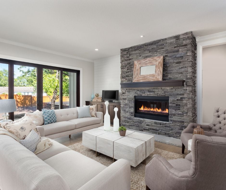 A bright, modern organic living room featuring a stone fireplace with a lit fire, neutral-toned sofas, a wooden coffee table, and large sliding glass doors leading to a backyard.
