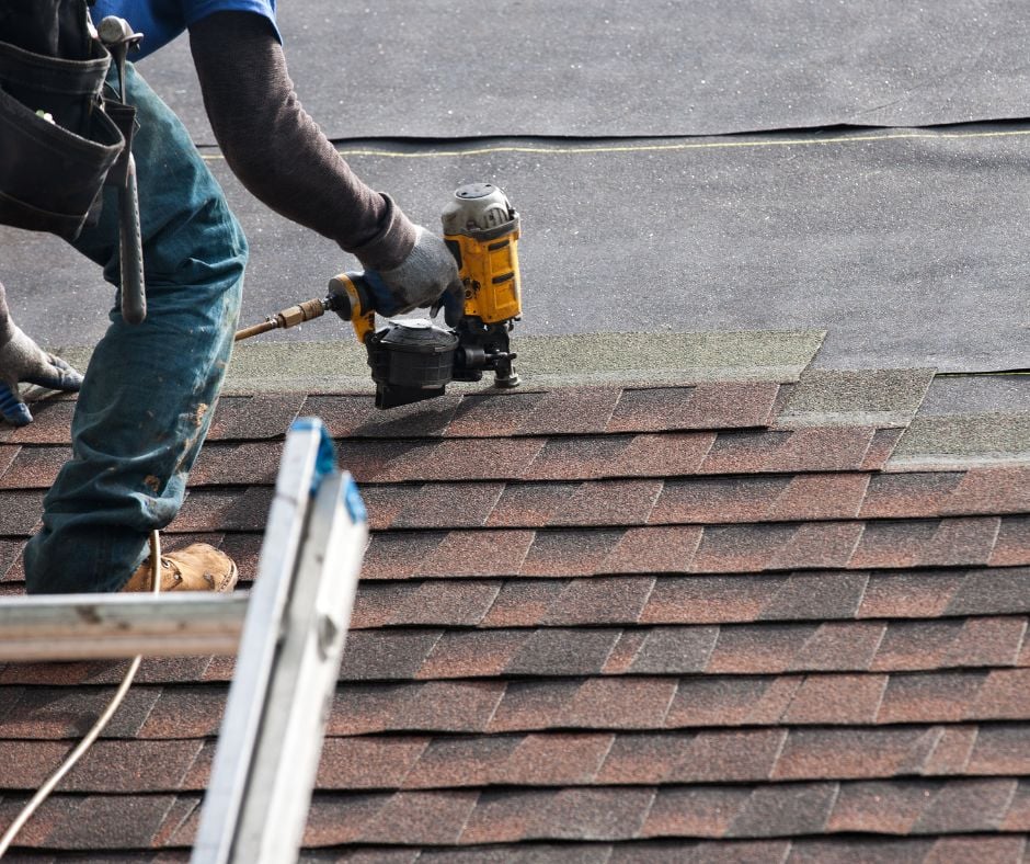 Close-up of a professional roofer installing new architectural asphalt shingles onto a residential roof using a yellow pneumatic nail gun.