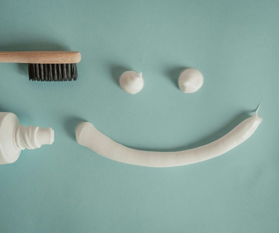 A minimalist flat lay on a light blue background featuring a wooden toothbrush with black bristles, an open tube of white toothpaste, and dollops of toothpaste arranged to form a smiling face.