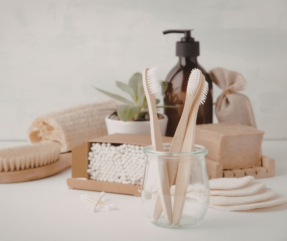 A collection of sustainable personal care items on a white surface, including bamboo toothbrushes in a small glass jar, a box of cotton swabs, a bar of natural soap on a wooden dish, reusable cotton rounds, and an amber glass pump bottle.