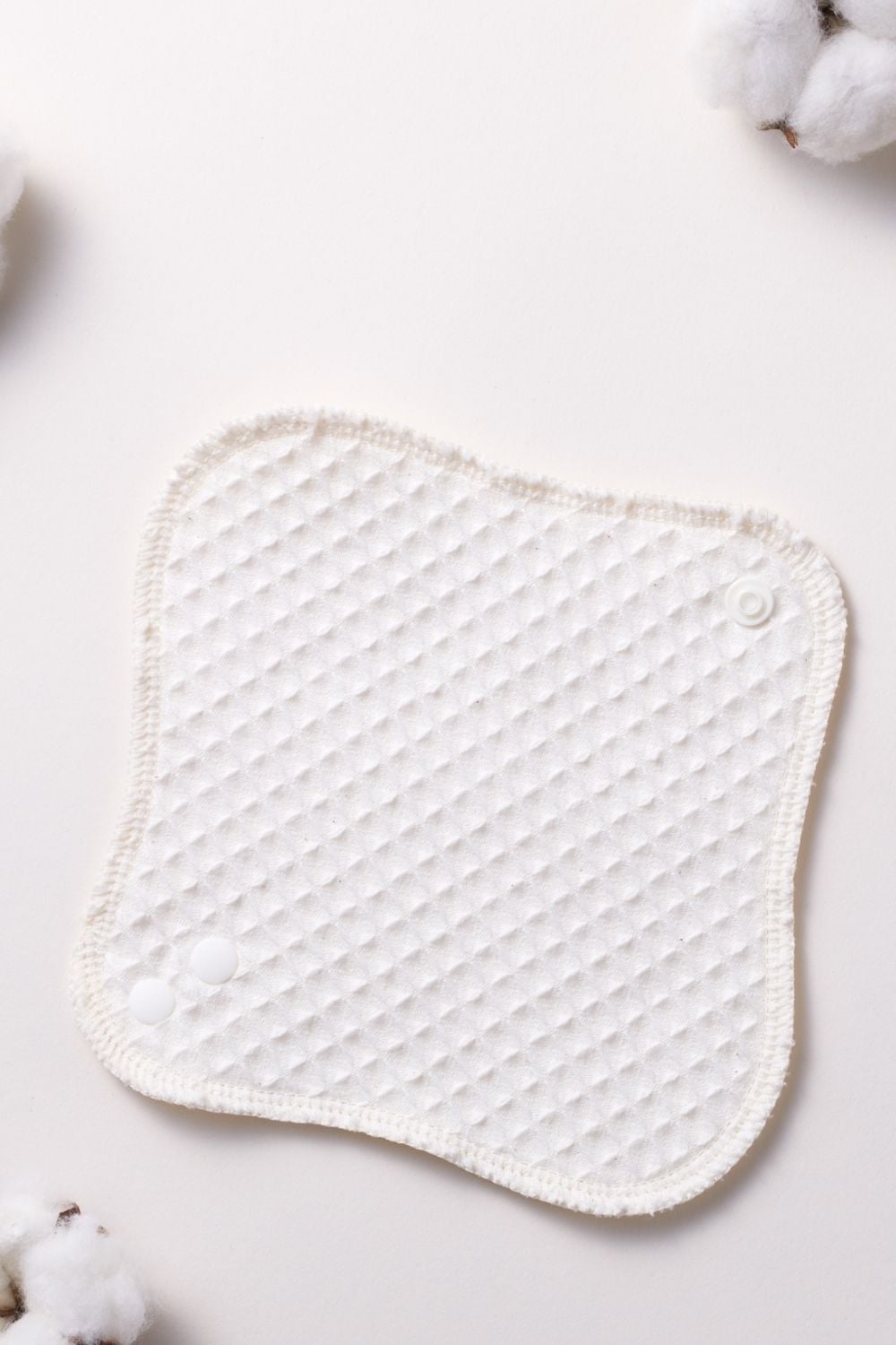 A close-up top view of a white, textured waffle-weave reusable organic cloth pad or makeup round with small white snap buttons, displayed on a clean white background with faint cotton blossoms in the corners.