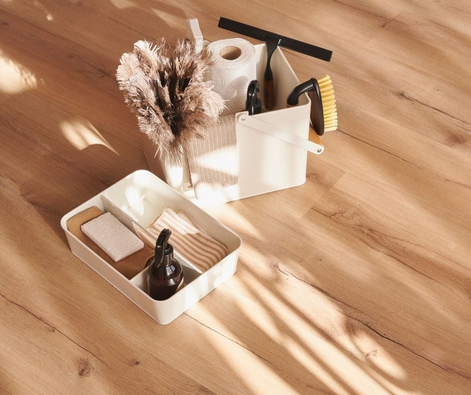 A high-angle shot of a minimalist white plastic cleaning caddy and a separate organizer tray sitting on light-colored wood flooring. The caddy is filled with neatly arranged eco-friendly cleaning tools, including a squeegee, brushes, a roll of paper, and an amber spray bottle.
