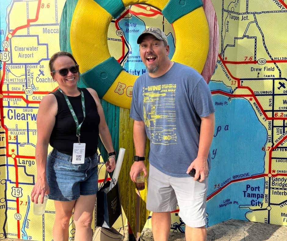 A man and woman smiling in front of a colorful map of Clearwater and Tampa with a large yellow life preserver frame.