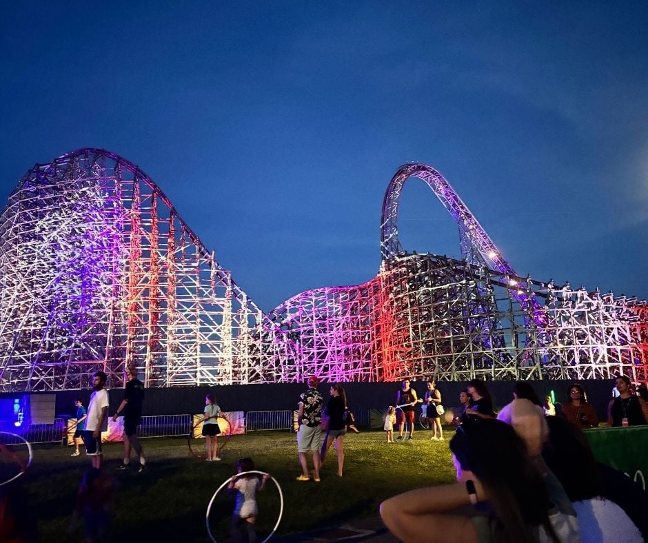 The Iron Gwazi wooden-steel hybrid roller coaster lit up with red, white, and blue lights at night while guests play with hula hoops in the foreground.