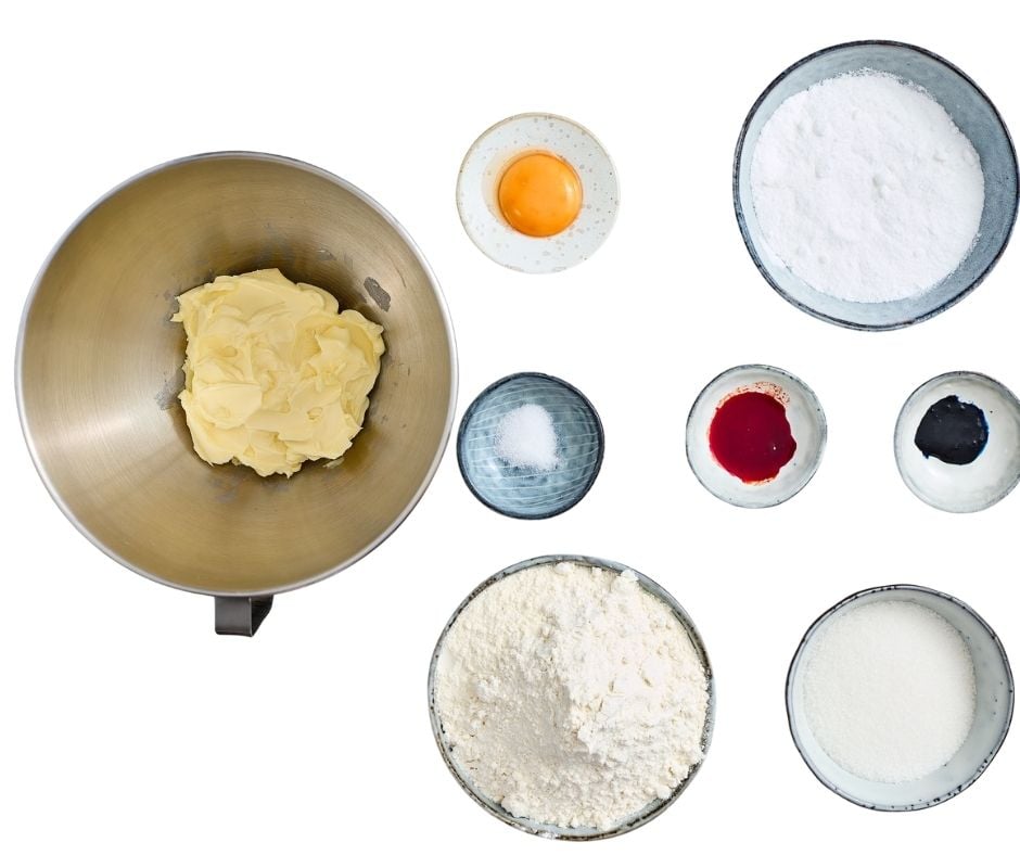 A flat-lay overhead view of ingredients, including a metal mixing bowl with butter, small speckled bowls holding a single egg yolk and granulated sugar, small ceramic bowls with salt, red gel food coloring, and blue gel food coloring, and two bowls of all-purpose flour, all arranged neatly on a white surface.