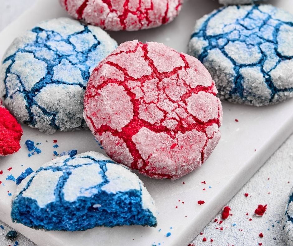 A close-up view focusing on several red and blue crinkle cookies spread on a white marble surface, including a prominent blue cookie piece split open to show its bright blue, moist interior and chewy texture, illustrating one of the "tips for success."